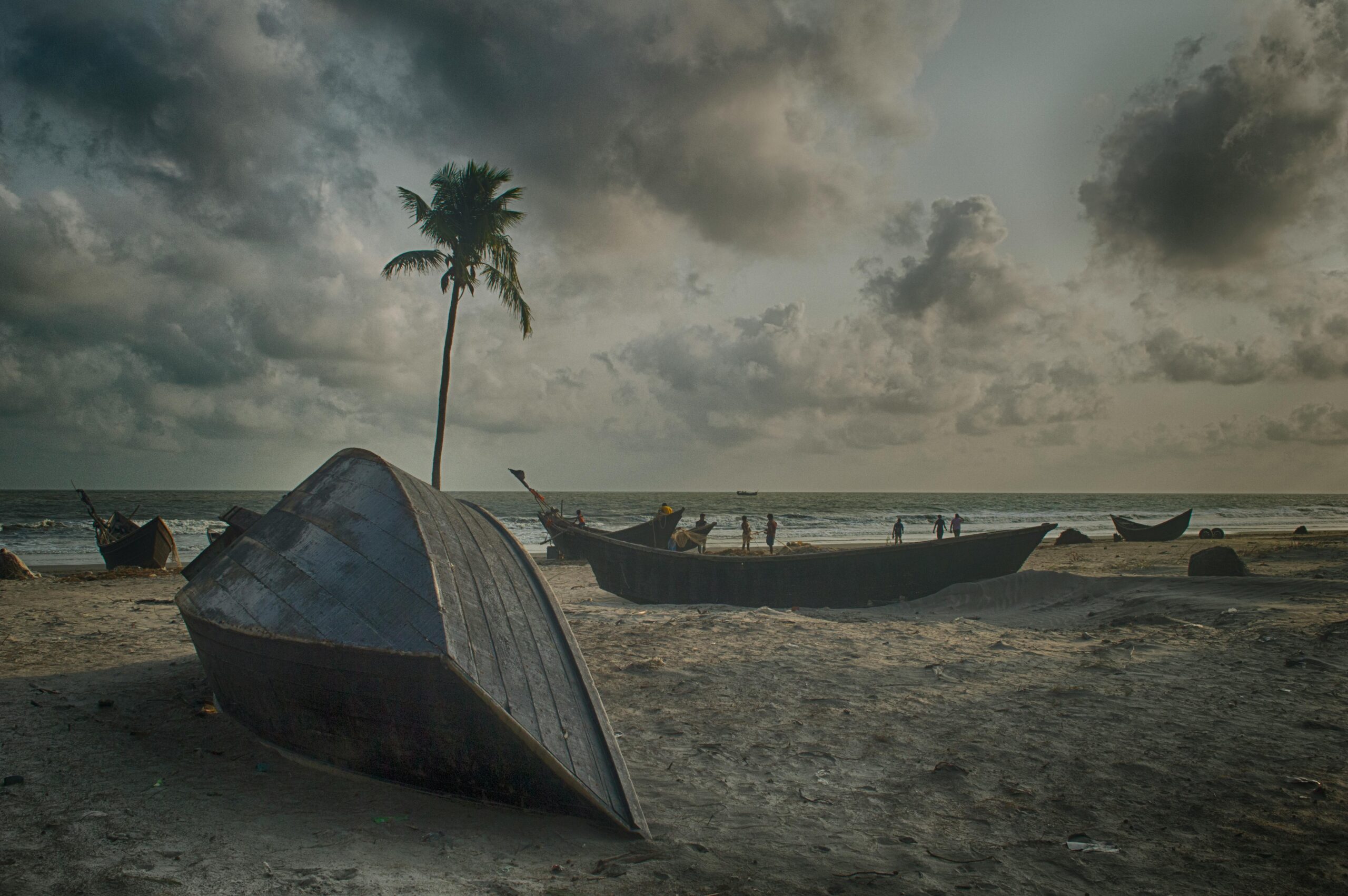Tranquil beach scene with wooden boats, palm tree, and cloudy sky in Cox's Bazar.